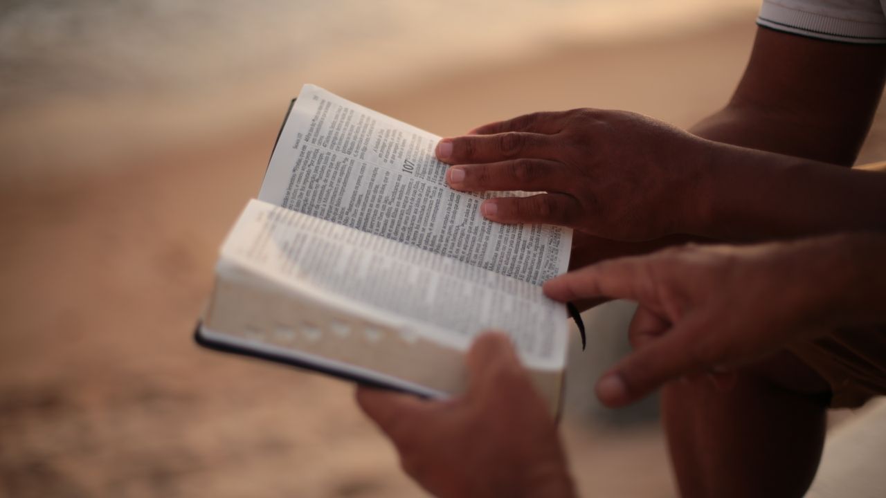 hero-gallery-04 Close-up of hands holding an open Bible on a beach, symbolizing faith and study.