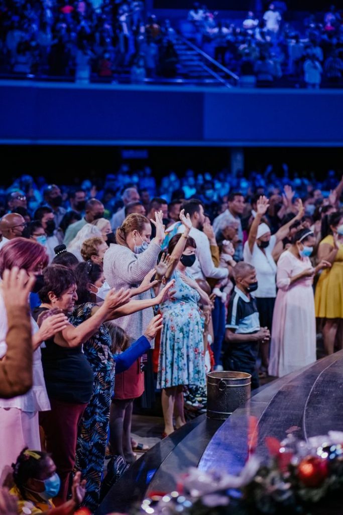 A large group of diverse people standing with arms raised, showcasing unity and diversity indoors.