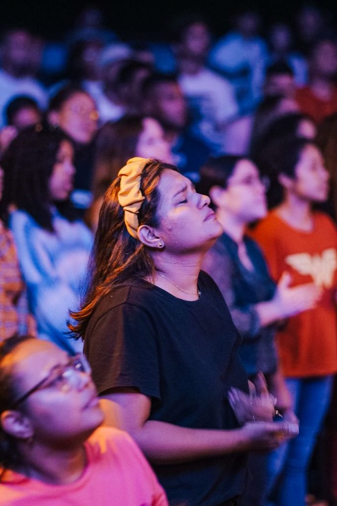 A woman in a crowd of diverse people praying with closed eyes and a serene expression.