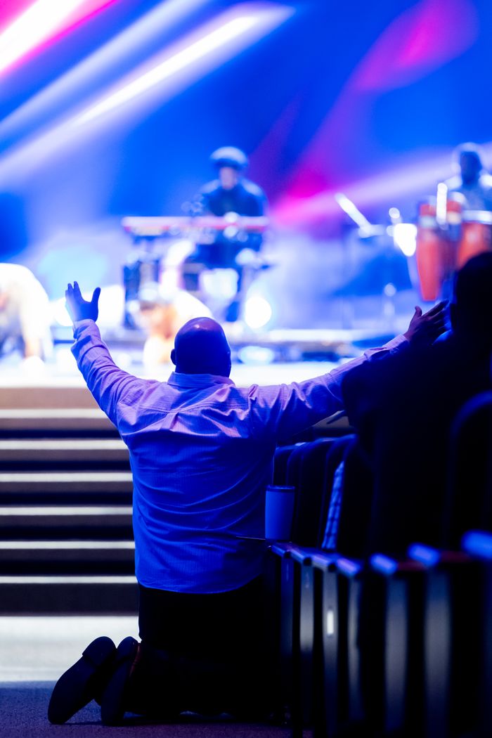 programs-01 A man kneels with arms raised during a live worship concert with vibrant lighting.