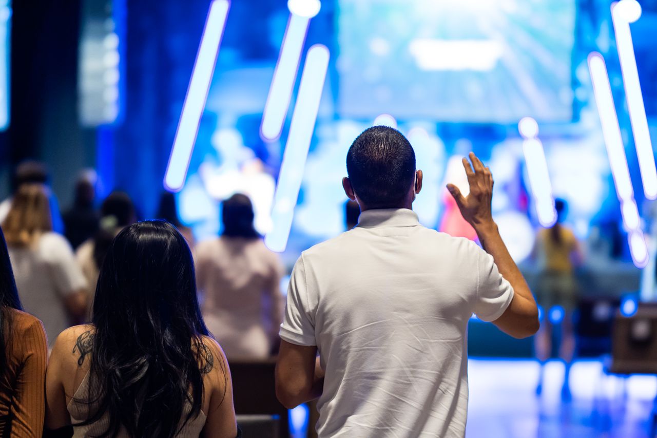 programs-02 People attending a worship service indoors, featuring a man raising his hand towards the stage.